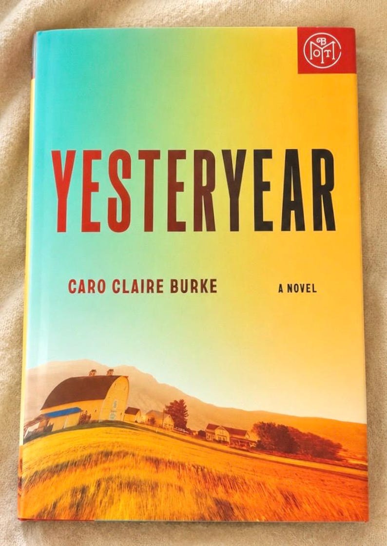 May include: A book titled Yesteryear by Caro Claire Burke with a cover featuring a gradient from teal to yellow. The cover displays a rural landscape with a yellow barn, fields, and mountains, and a red logo in the top right corner.