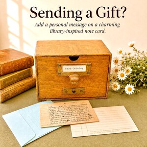 May include: A wooden card catalogue drawer with a brass pull and label holder reading Card Catalog. Beside it are three stacked books, a light blue envelope, a handwritten index card, a lined note card, and a bouquet of white daisies.