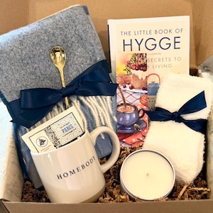 May include: A gift box containing a blue woven throw blanket with a gold spoon, a white mug with the text HOMEBODY, a copy of The Little Book of Hygge, a white fuzzy cloth, and a round candle in a tin, all tied with dark blue satin ribbons.