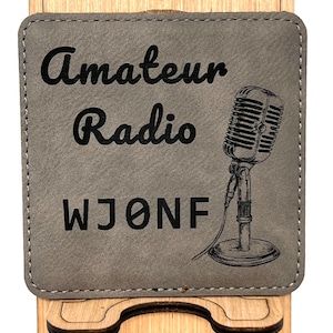 May include: A grey faux leather phone stand with a black engraved design. The design includes the words "Amateur Radio" and "WJONF" with a vintage microphone illustration.