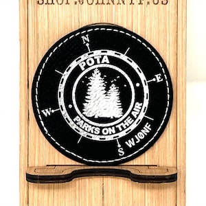 Pode incluir: Uma caixa de madeira com um círculo de couro preto no estilo de bússola com o texto "POTA Parks on the Air" e uma silhueta branca de duas árvores de pinho. A caixa tem uma pequena prateleira na parte inferior.