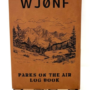 Puede incluir: Un diario encuadernado en cuero marrón con el texto "WJØNF PARKS ON THE AIR LOG BOOK" y una ilustración en blanco y negro de una cordillera con árboles.