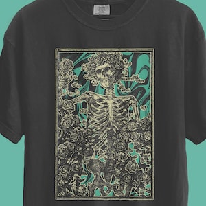 May include: A black t-shirt with a graphic of a skeleton surrounded by roses and teal accents. The design is centered on the front of the shirt. The shirt is hanging on a wooden hanger against a teal background.