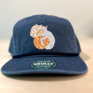 May include: A navy blue baseball cap with a dark blue rope trim across the base of the crown. The front features a graphic of an orange tabby cat and a light blue cat hugging, with the text CUDDLES FOR WORLD PEACE and a peace sign symbol.