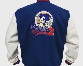 May include: A blue varsity jacket with white sleeves and red and blue striped ribbed trim at the collar, cuffs, and hem. The back features a large embroidered patch of Sonic the Hedgehog with the text Sonic The Hedgehog 2 in blue and red.