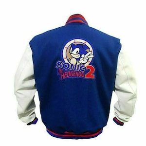 Puede incluir: Chaqueta de béisbol azul y blanca con un logotipo de Sonic the Hedgehog 2 en la espalda. El logotipo presenta a Sonic the Hedgehog en un círculo rojo con estrellas blancas.