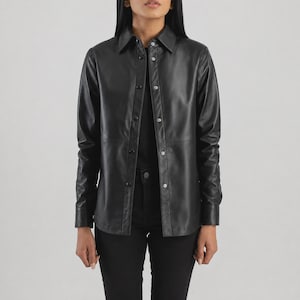 May include: A black leather shirt featuring a classic collar, long sleeves with buttoned cuffs, and a front button-down closure. The garment has a smooth finish, a straight hemline, and visible seams across the chest and torso, providing a structured look.