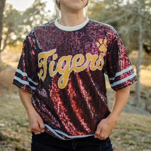 May include: A red sequined shirt with white and blue stripes on the sleeves. The shirt has the word "Tigers" written in gold letters across the front.