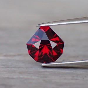 May include: A faceted, deep red gemstone with a shield-like pentagonal shape. The stone features precise geometric cuts that create a symmetrical pattern of light and dark reflections across its surface, held by a pair of silver-toned metal tweezers.