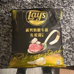 Puede incluir: Una bolsa de patatas fritas Lay's con fondo negro y detalles dorados. La bolsa presenta una imagen de un filete de ribeye con una trufa encima. El texto de la bolsa dice "Lay's" y "Ribeye Steak".