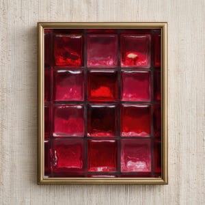 以下が含まれることがあります： 赤いガラスブロックのグリッドが特徴の額装されたアートワーク。ブロックは、濃いクリムゾンから明るい赤色まで、さまざまな色合いがあります。フレームは細く金色で、アートワークはテクスチャ加工されたオフホワイトの背景に表示されています。