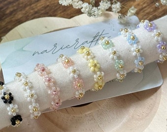 May include: A collection of beaded daisy rings in various colors, including black, white, pink, yellow, green, blue, and purple. Each ring features small gold beads and is displayed on a white cloth. The rings are handmade and delicate.