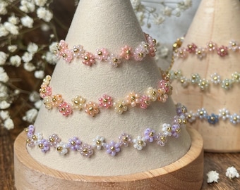 May include: Close-up of several beaded bracelets, each featuring delicate floral designs. The bracelets are displayed on wooden cone-shaped stands. The color palette includes soft pastels like pink, peach, lavender, and blue, with gold accents.