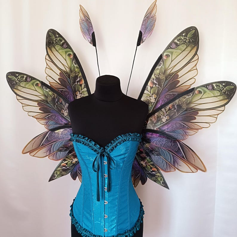 K&ouml;nnte beinhalten: Ein Paar gro&szlig;e, kunstvoll gestaltete Feenfl&uuml;gel mit einem blaugr&uuml;nen Korsett. Die Fl&uuml;gel bestehen aus durchsichtigem Stoff mit einem floralen und blattf&ouml;rmigen Muster in Gr&uuml;n-, Lila- und Braunt&ouml;nen. Das Korsett hat einen Spitzenbesatz und eine Satin-Oberfl&auml;che.