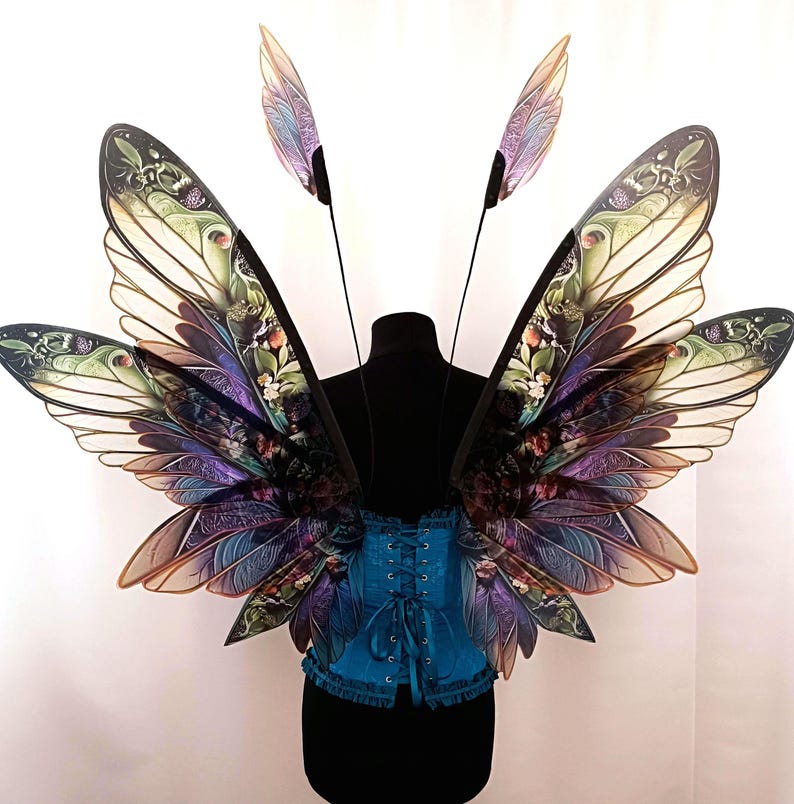 K&ouml;nnte beinhalten: Ein Paar gro&szlig;e, schillernde Schmetterlingsfl&uuml;gel mit einem blaugr&uuml;nen Korsett. Die Fl&uuml;gel sind mit floralen und Blattmotiven verziert und haben eine durchscheinende, fast glasartige Optik.