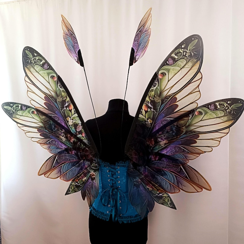 K&ouml;nnte beinhalten: Ein Paar gro&szlig;e, schillernde Schmetterlingsfl&uuml;gel mit filigranen Details und einem Farbspiel aus Gr&uuml;n, Lila und Gold. Die Fl&uuml;gel sind an einem schwarzen Mannequin-Torso befestigt, der ein blaues Korsett tr&auml;gt.
