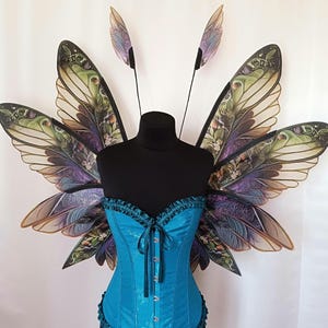 K&ouml;nnte beinhalten: Ein Paar gro&szlig;e, kunstvoll gestaltete Feenfl&uuml;gel in Teal- und Lilat&ouml;nen. Die Fl&uuml;gel bestehen aus durchsichtigem Material mit einem floralen Muster und sind an einem schwarzen Mannequin befestigt.