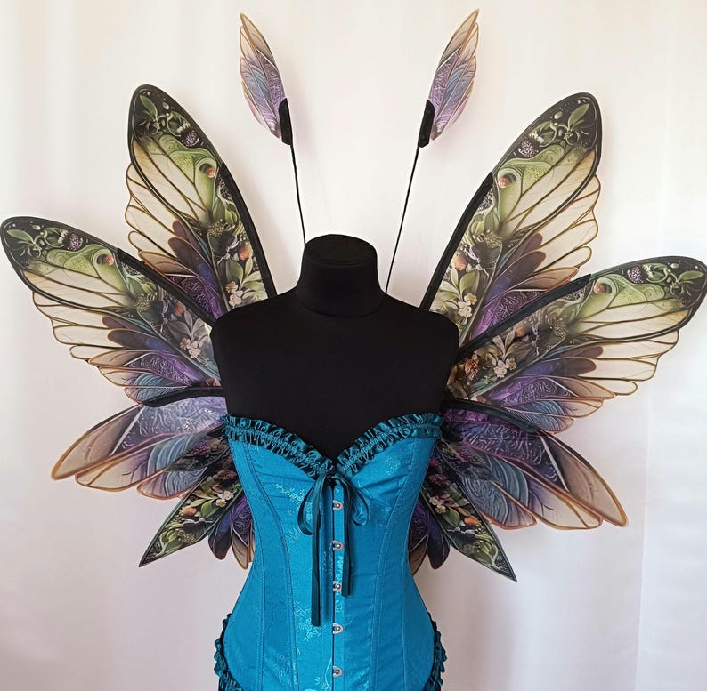K&ouml;nnte beinhalten: Ein Paar gro&szlig;e, kunstvoll gestaltete Feenfl&uuml;gel in Teal- und Lilat&ouml;nen. Die Fl&uuml;gel bestehen aus durchsichtigem Material mit einem floralen Muster und sind an einem schwarzen Mannequin befestigt.