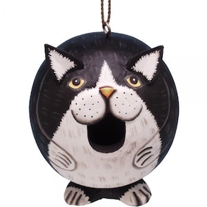 May include: Black and white cat-shaped birdhouse with a round body and a large opening for birds to enter. The birdhouse has a yellow nose and yellow eyes.
