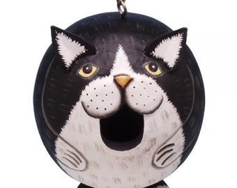 May include: Black and white cat-shaped birdhouse with a round body and a large opening for birds to enter. The birdhouse has a yellow nose and yellow eyes.