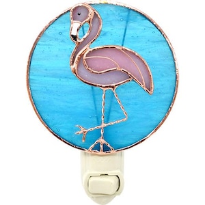 May include: A stained glass night light featuring a pink flamingo standing on a blue background. The night light is made of copper and glass and has a white plug-in base.