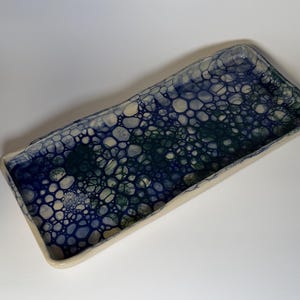 Könnte beinhalten: Rechteckige Keramikschale mit blauem, grünem und weißem Blasenmuster. Die Schale hat einen erhöhten Rand und einen cremefarbenen Rand. Der Innenraum weist ein strukturiertes, organisches Design auf, das sich zum Servieren oder Ausstellen eignet.
