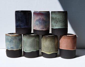 May include: Seven cylindrical ceramic cups arranged in two rows. Each cup features a matte black base and a unique reactive glaze top in shades of deep blue, pink, sage green, light blue, mint, olive, and terracotta, creating a varied color palette.