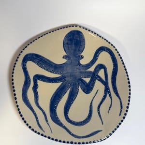 Könnte beinhalten: Keramikplatte mit handgemaltem blauen Oktopus-Design. Der Oktopus ist mittig auf der cremefarbenen Platte platziert, mit seinen Tentakeln, die sich ausbreiten. Der Rand der Platte ist mit kleinen blauen Punkten verziert, die einen Rand bilden. Für Küchen- oder Esszimmerdekoration.