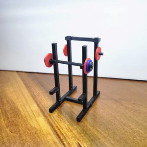 Peut inclure: Un petit rack d'haltérophilie noir avec une barre et des poids. La barre a des poids rouges et bleus à chaque extrémité. Le rack est en plastique noir et est posé sur une surface en bois.