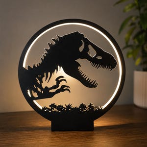 May include: Black dinosaur silhouette lamp with a circular design. The lamp features a T-Rex design within a glowing white circle. The base is rectangular, and the lamp is placed on a wooden surface. The background is neutral.