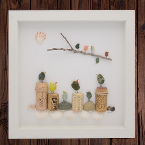 May include: A framed art piece featuring a whimsical arrangement of small, colorful birds crafted from pebbles and stones. The birds perch on corks and stones, with a branch overhead. Seashells are at the base, creating a nature-inspired scene.