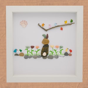 May include: Framed pebble art depicting a bunny and birds. The bunny is constructed from stacked pebbles, with a flower garden in front. Colourful birds perch on a branch above. A seashell is in the upper left corner.