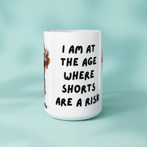 Mockup showing an 11oz or 15oz mug with a sublimation design depicting a humorous old man and the text I am at the age where shorts are a risk Set against a light blue background to illustrate how the design appears on a physical mug