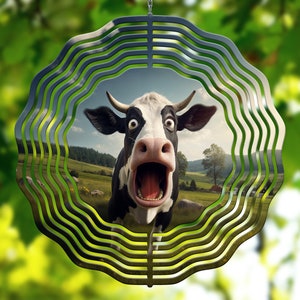 May include: A metal wind spinner with a black and white cow face in the center. The cow is looking directly at the camera with its mouth open and tongue sticking out. The background is a blurry image of a green field and blue sky.