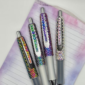May include: Four pens with iridescent rhinestones. The pens have silver accents and gray grips. The rhinestones are arranged in rows, creating a sparkling effect. The pens are resting on a lined notepad with a watercolor design.