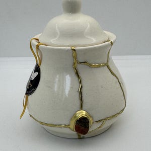 Puede incluir: Un tarro de cerámica color crema con tapa, con reparaciones kintsugi doradas. El tarro tiene forma redonda con un pequeño pomo en la parte superior y un pequeño acento de piedra marrón rojizo. Una etiqueta con texto cuelga del tarro.