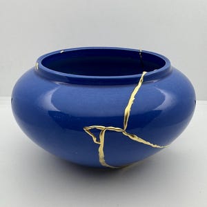 May include: A blue ceramic bowl with a wide opening and rounded body. The bowl has been repaired with gold-colored kintsugi, highlighting the cracks. The bowl is a decorative item.
