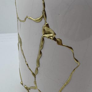 May include: A white ceramic vase with gold-colored kintsugi-style repairs. The gold lines trace the cracks and breaks in the vase, creating a unique and artistic design. The vase has a smooth, glossy finish.