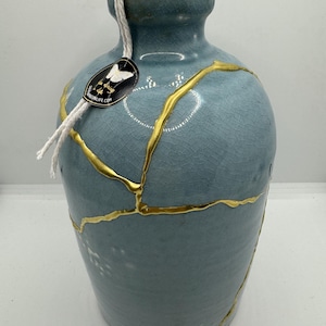 May include: A light blue ceramic vase with gold crackle glaze and a cork stopper. The vase has a white string tied around the neck and a black label with a butterfly design and the text "The Butterfly Life Co."