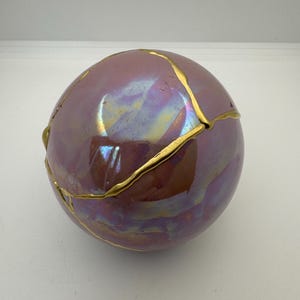 May include: A spherical, iridescent ceramic object with a purple and gold color scheme. The surface has a glossy finish and is adorned with gold-colored accents that highlight the cracks, creating a unique aesthetic.