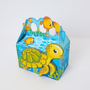 Peut inclure: Une boîte à dragées en carton bleu et jaune avec un motif de tortue et de poissons. La boîte a une poignée et porte l'inscription "Pedro" sur le côté.
