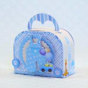 Puede incluir: Caja de regalo para baby shower de color azul y blanco con asa. La caja presenta un mono estampado azul y blanco, un coche azul y globos azules y blancos. La caja está decorada con huellas de patas.