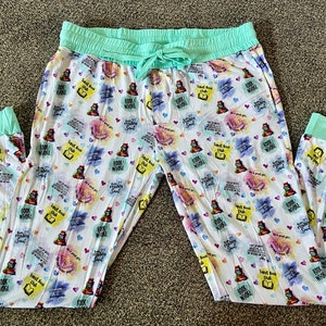 May include: White pajama pants with a mint green waistband and cuffs. The pants have a colorful print of stacked books, hearts, and the text "Sweet Book Club" and "Books Whole World".