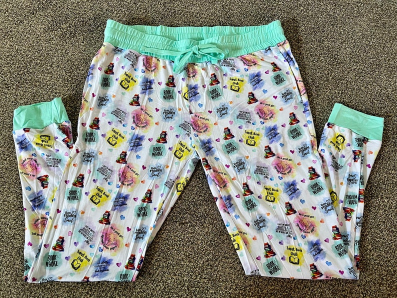 May include: White pajama pants with a mint green waistband and cuffs. The pants have a colorful print of stacked books, hearts, and the text "Sweet Book Club" and "Books Whole World".