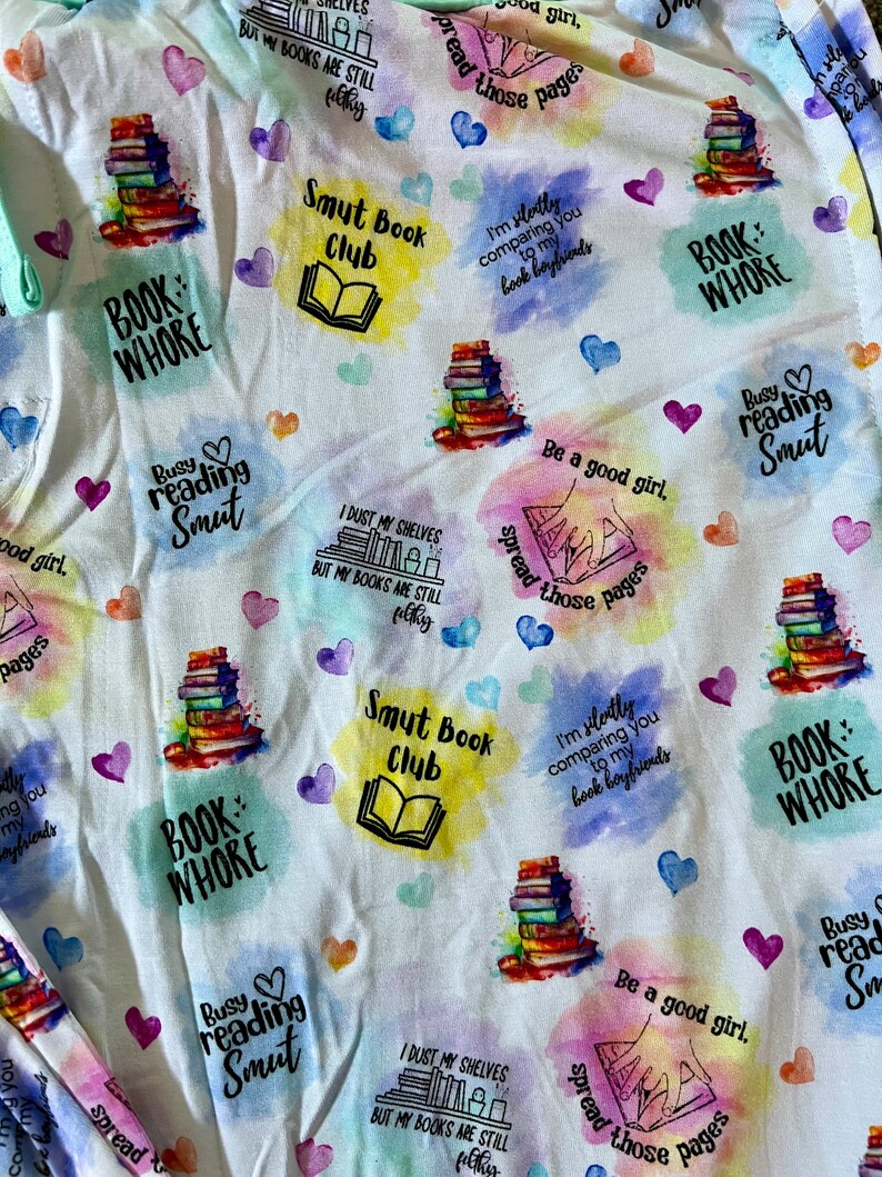 May include: White fabric with a colorful, watercolor-style pattern featuring stacks of books, hearts, and text. The text includes phrases like "Book Whore", "Smut Book Club", "Busy Reading Smut", and "I dust my shelves but my books are still filthy".