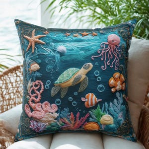 May include: Decorative square pillow featuring an underwater scene with a sea turtle, starfish, jellyfish, and coral. The pillow has a dark teal background with embroidered details and a gold border. The pillow is placed on a white cushion.
