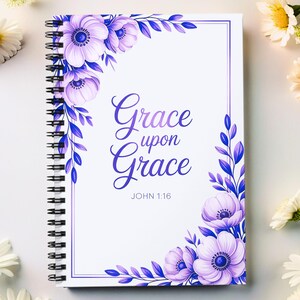 May include: A spiral-bound notebook with a white cover featuring the words "Grace upon Grace" in a purple script, surrounded by purple floral and leaf designs. The text "John 1:16" is below the main text. Daisies and other white flowers are arranged around the notebook.