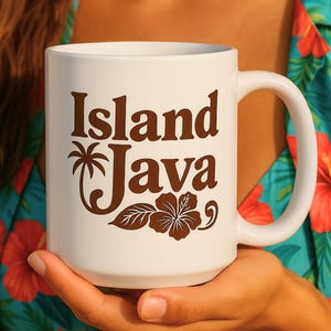 Könnte beinhalten: Weiße Keramik-Kaffeetasse mit einer braunen Grafik, auf der "Island Java" mit einer Palme und einer Hibiskusblüte steht.