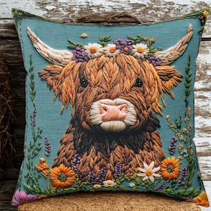 May include: A decorative square pillow featuring a detailed embroidered image of a Highland cow. The cow has a brown and tan coat, a pink nose, and is adorned with a floral crown. The pillow has a teal background and is decorated with flowers.