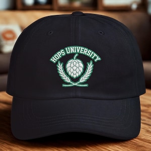 Può includere: Cappellino da baseball nero con la scritta "HOPS UNIVERSITY" in verde e bianco. Il logo presenta un cono di luppolo e spighe di grano. Il cappellino è su una superficie di legno.